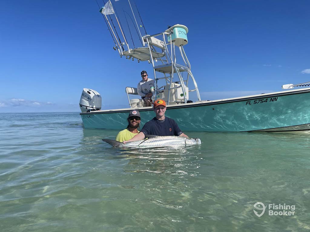 Two anglers standing just above waist-deep in the shallow waters of Tampa Bay, holding a Tarpon fish, while a center console charter fishing boat and its captain is stationary behind them on a clear day