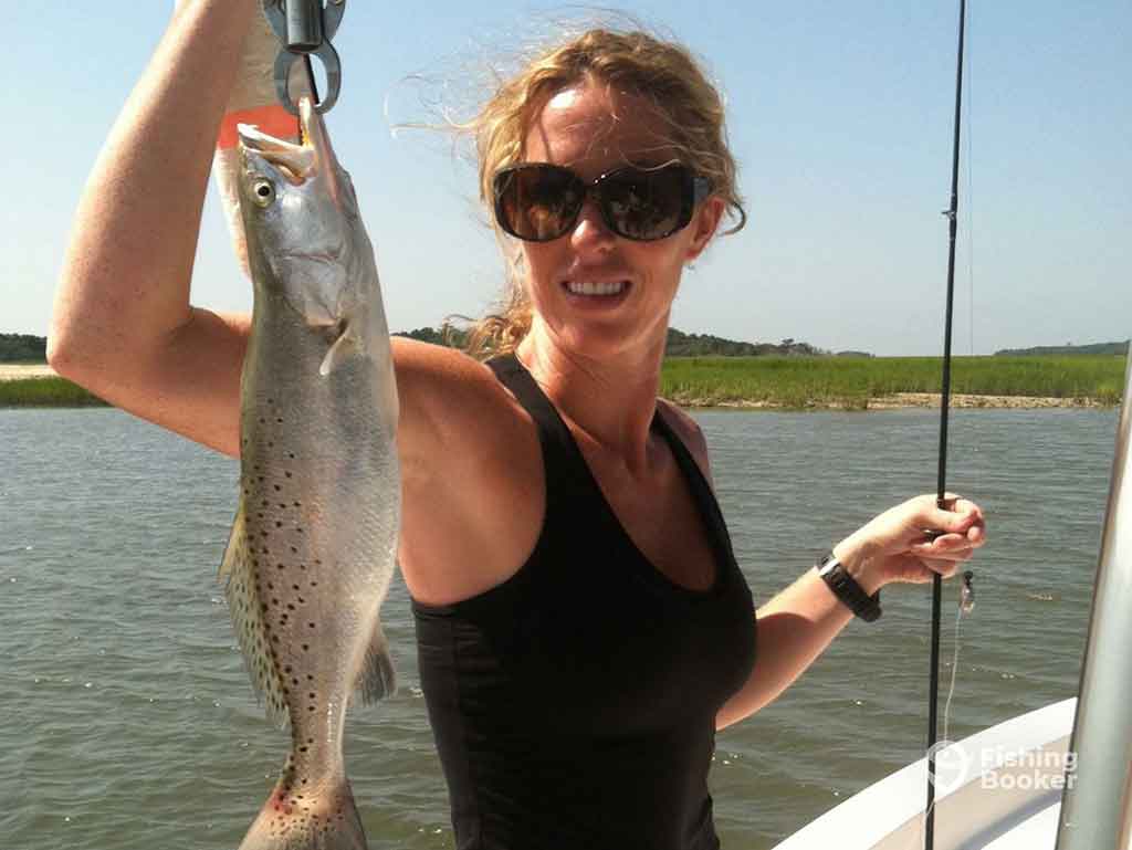 A woman in sunglasses, standing on a fishing boat in the inshore waters near Kiawah Island, SC, and holding up a Spotted Seatrout vertically with one hand while holding a fishing rod in the other