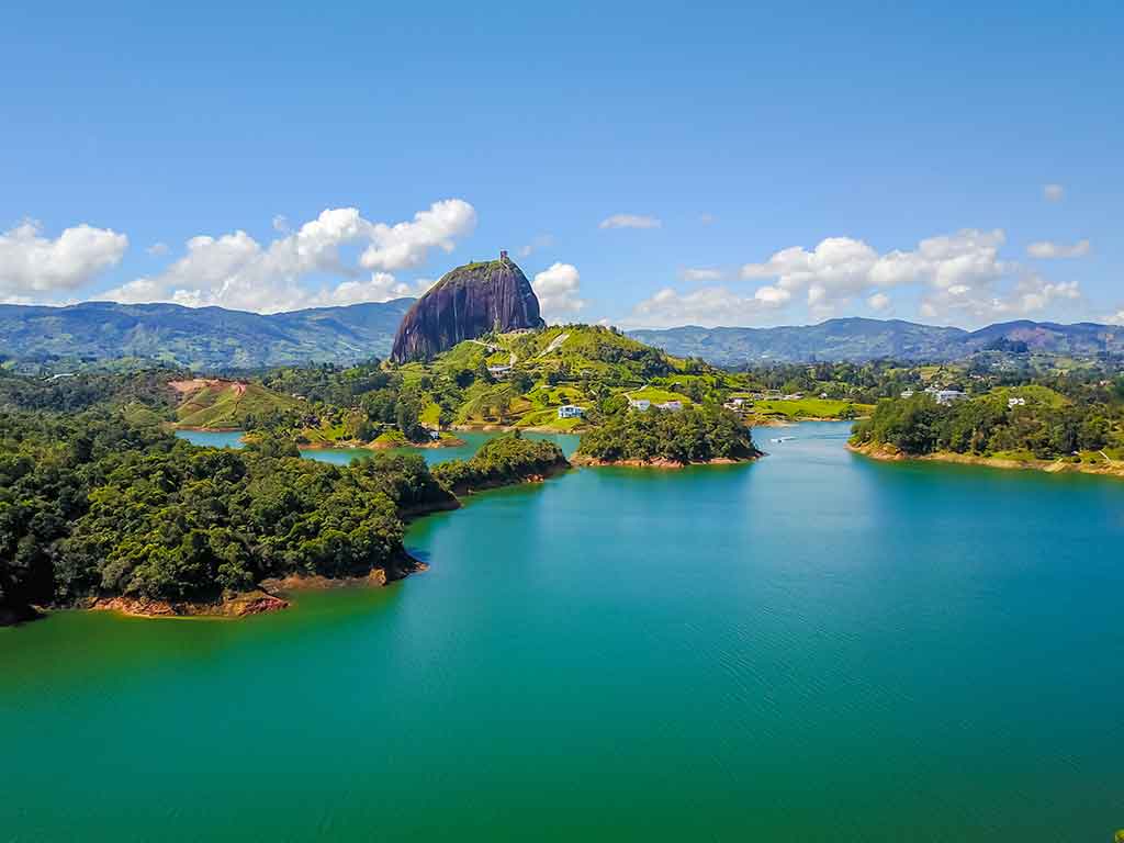 An aerial view of a lake in Colombia with crystal clear blue waters dominating the image, giving way to lush green hills and one rounded peak in the center of the image on a clear day
