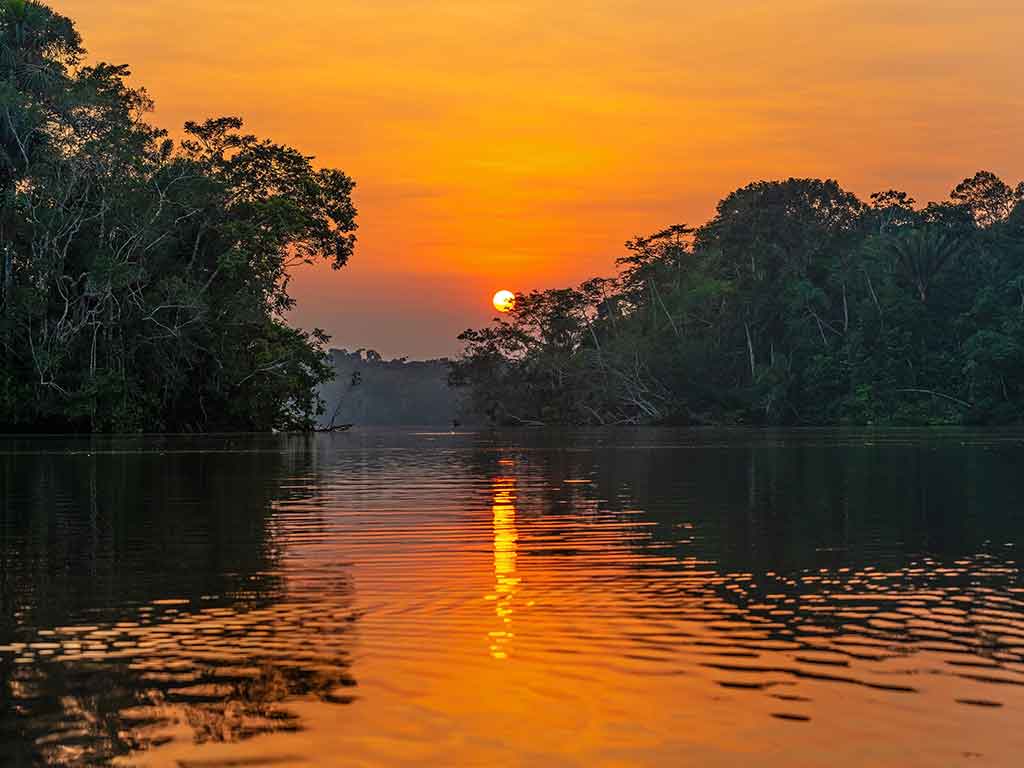 A view across the river towards a setting sun in the middle of the image, creating an orange hue, with trees visible on either side of the calm waterway
