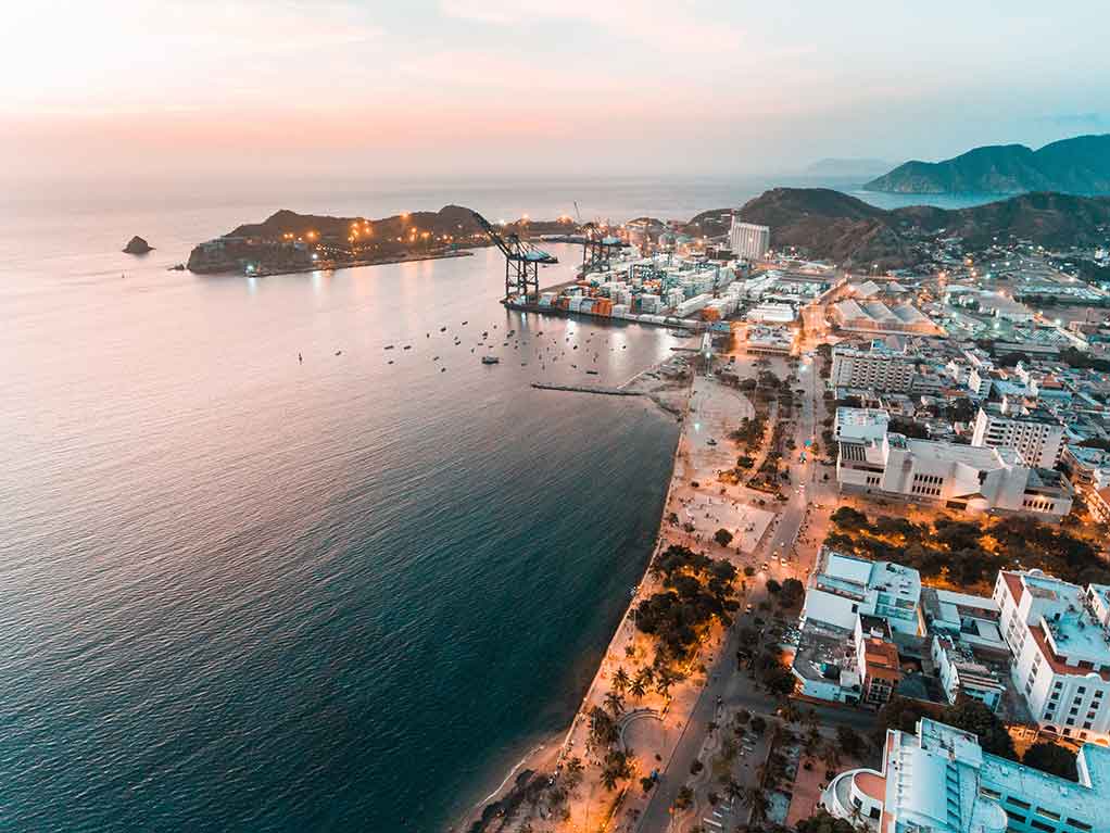 An aerial view of Santa Marta, Colombia, at sunset, with a port visible in the distance and the busy city on the right of the image contrasting with the calm waters of the ocean on the left