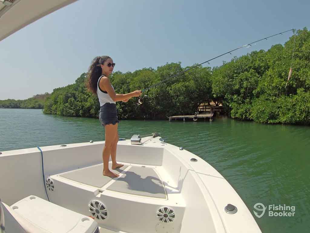 A woman standing on the bow of a fishing boat in the mangroves of Colombia, casting a fishing line into the water on a clear day