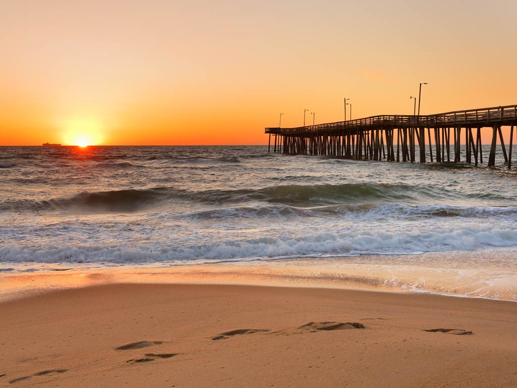 A sunset photo of the fishing pier in Virginia Beach, as small waves roll towards the pristine sandy beach