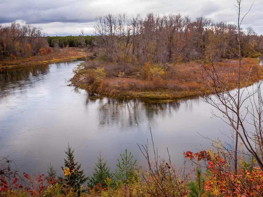 A photo of the Au Sable River near Grayling, Michigan during late fall, with only a few leaves left on the trees under cloudy skies.