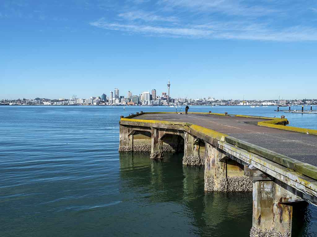 A view across a fishing pier towards a bay separating it from the city centre of Auckland, with a solitary angler visible at the end of the pier and the skyline of the city visible in the distance on a clear day