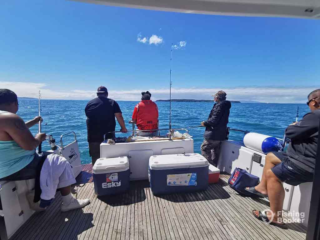 A view out the back of a fishing charter in Auckland, with five people standing around and one trolling line trailing behind the boat on a clear day in the deep blue waters