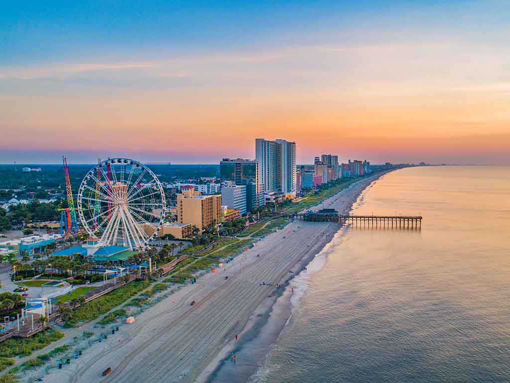 An aerial view of Myrtle Beach, SC, with a beach dominating the landscape, a ferris wheel in the foreground, and fishing pier visible in the distance at sunset