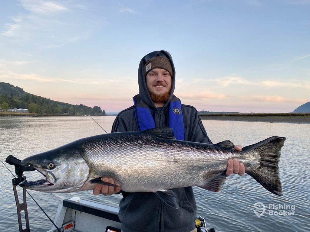 A man in winter gear standing on a fishing charter and holding a large Chinook Salmon aboard a boat in Oregon at sunset on a clear day