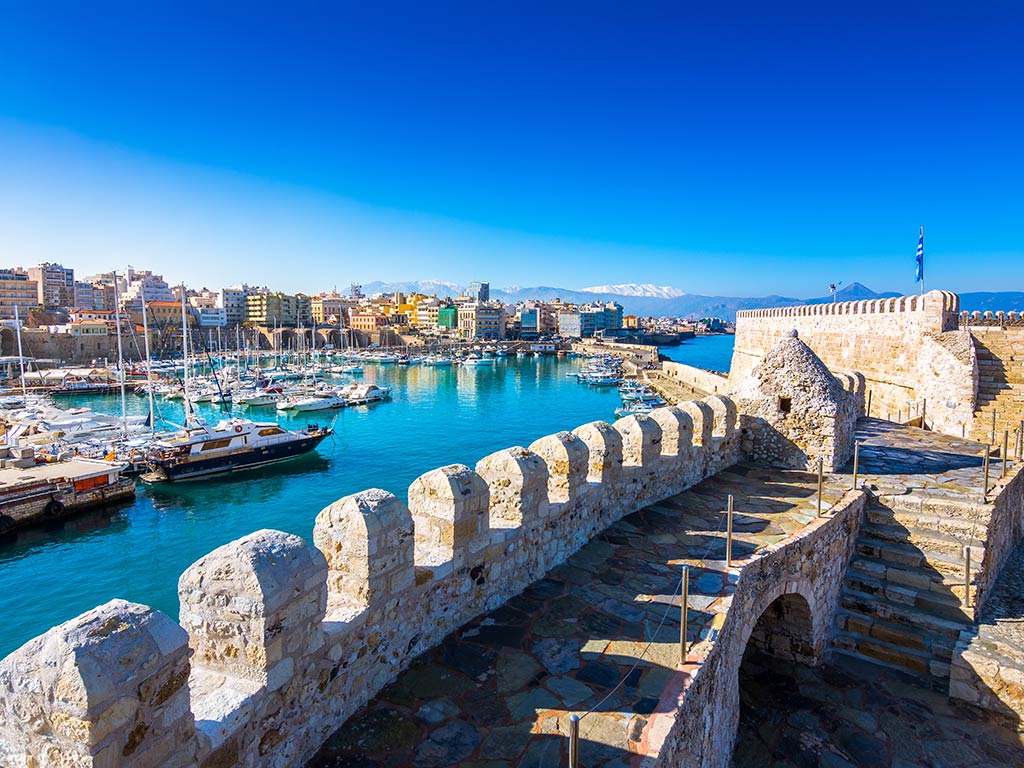 A view from atop the fortress walls of a marina in Crete, full of fishing boats, with the ramparts of the fortress visible in the foreground on a clear day