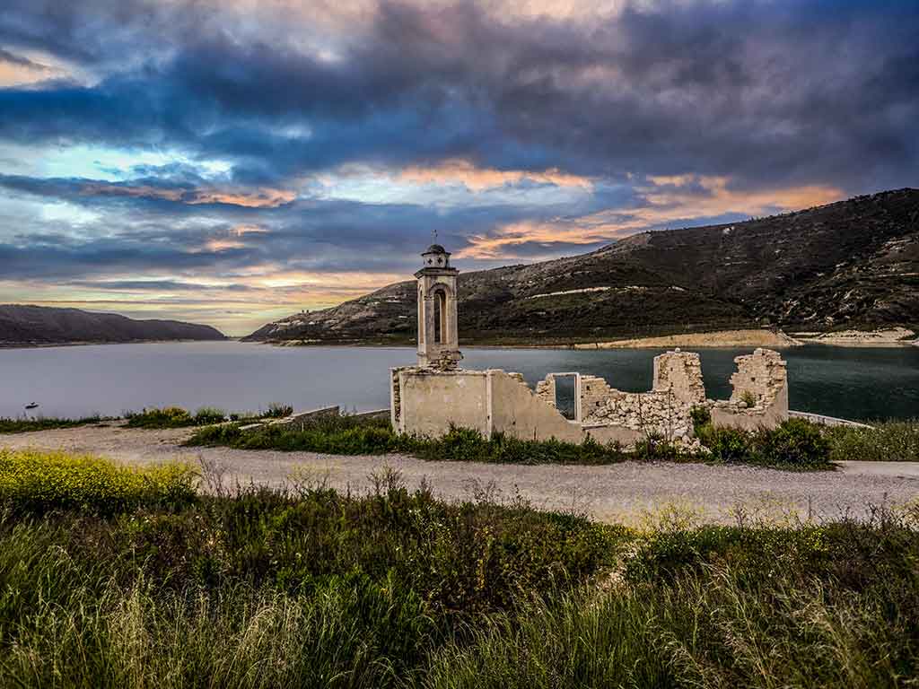 A view of a ruined church in Kouris Dam, Cyprus, with a lake extending behind it and mountains visible either side on a cloudy evening