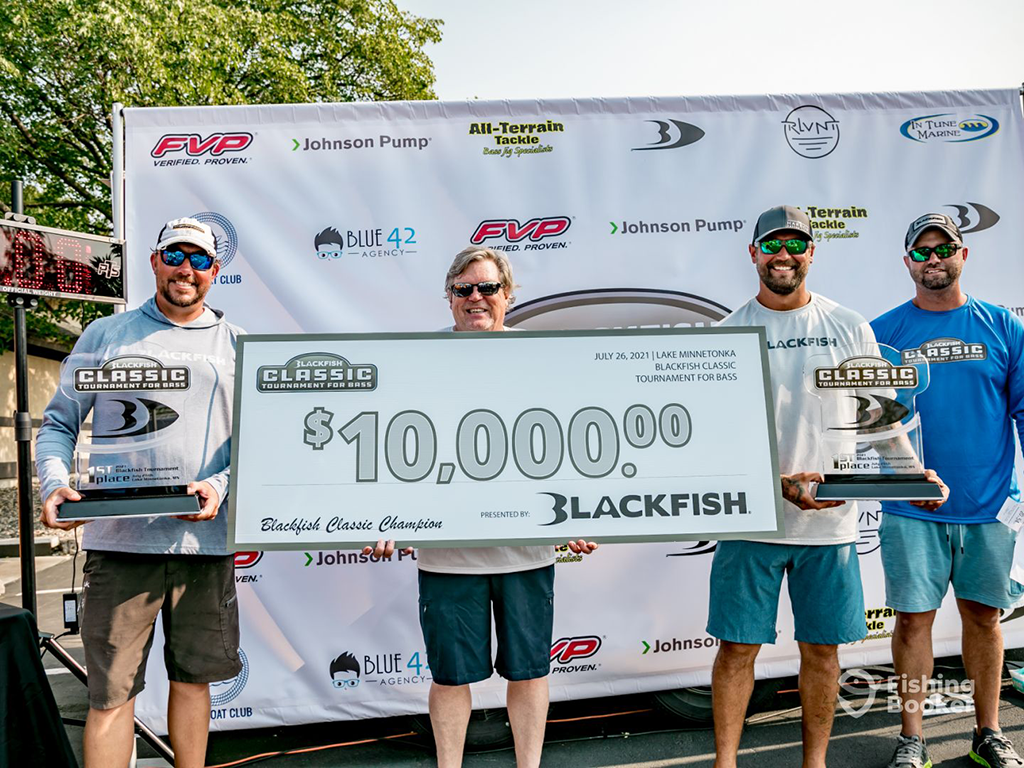 A man stands in between three men in front of a board full of advertisements holding a large check for $10,000 won in a Bass fishing competition in Minnesota.