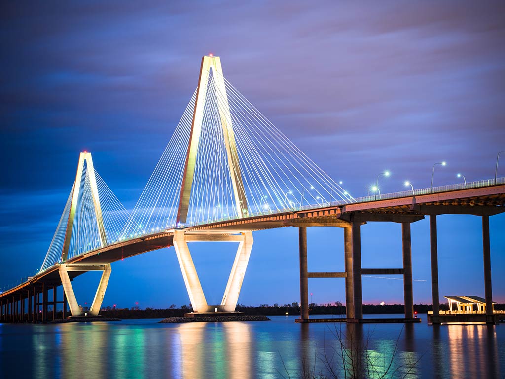 A view from the water towards the Arthur Ravenel Jr. Bridge, towering over the Cooper River at night, with the lights of the bridge reflected on the calm water