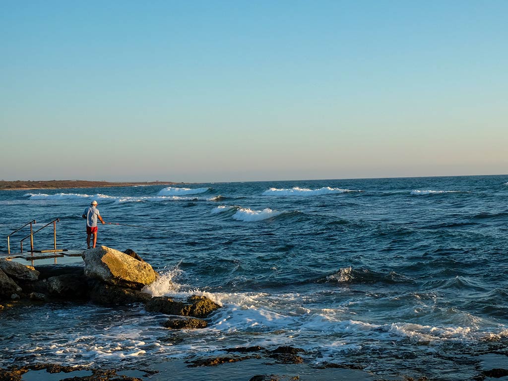 A view from shore in Paphos, Cymrus, looking towards waves crashing into the land and a jetty sticking out on the left of the image with a lone angler on it