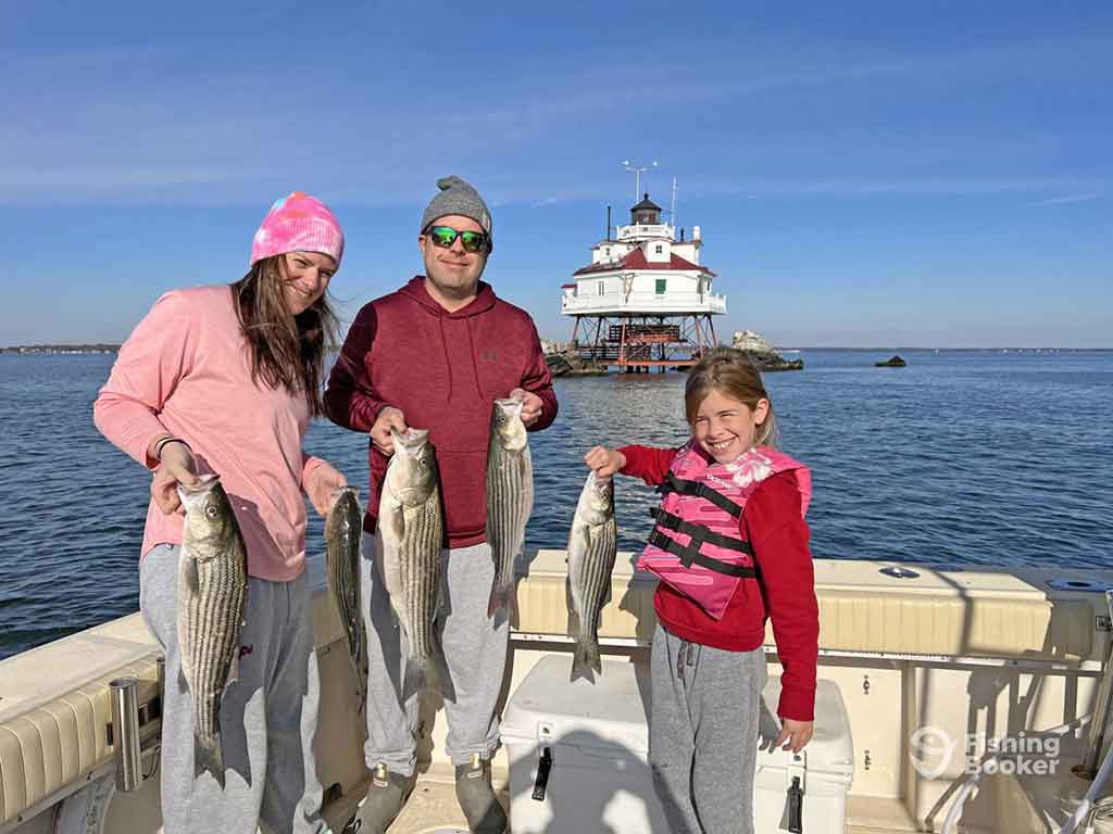 Two adults and a young girl aboard a fishing charter in Delaware, each holding up at least one Striped Bass with a lighthouse visible across the water behind them on a clear day