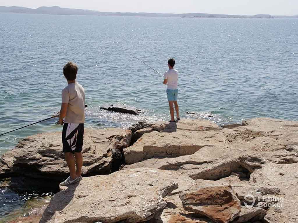 Two anglers standing on rocks on the shore of Lake Nasser casting their lines into the water on a clear day