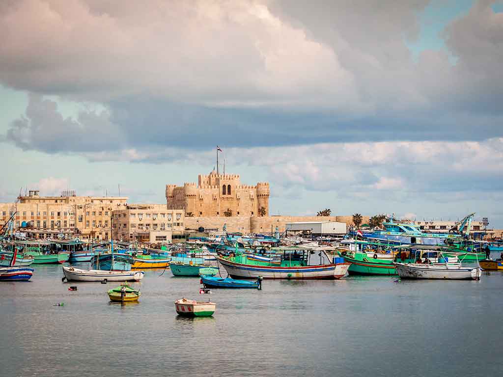 A view across the water towards the fortress in Alexandria on Egypt's Mediterranean coast with numerous boats visible in front of it on a cloudy day