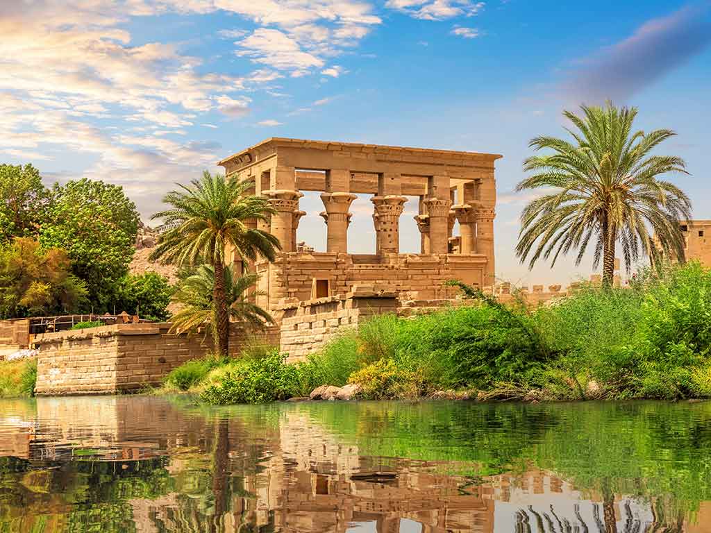 A view of Philae Temple in Egypt with greenery and palm trees visible in the foreground on a clear day
