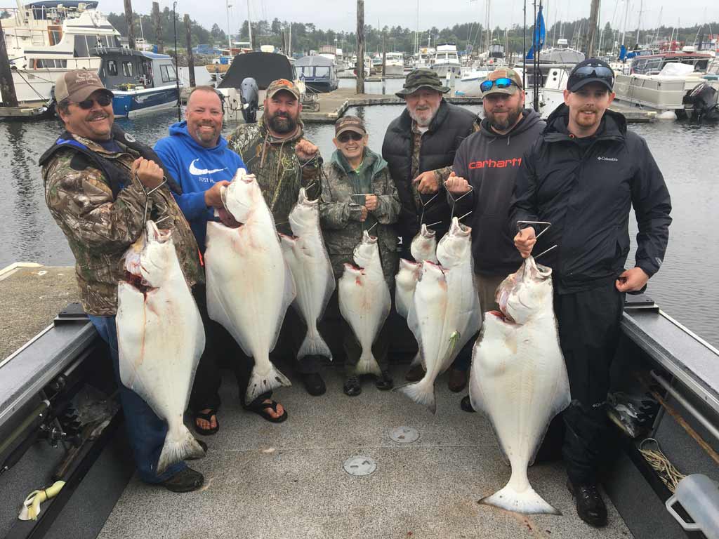 A group of anglers posing on the deck of a charter boat in Oregon, holding six Halibut they caught fishing during the all-depth season, with other docked boats visible behind them.