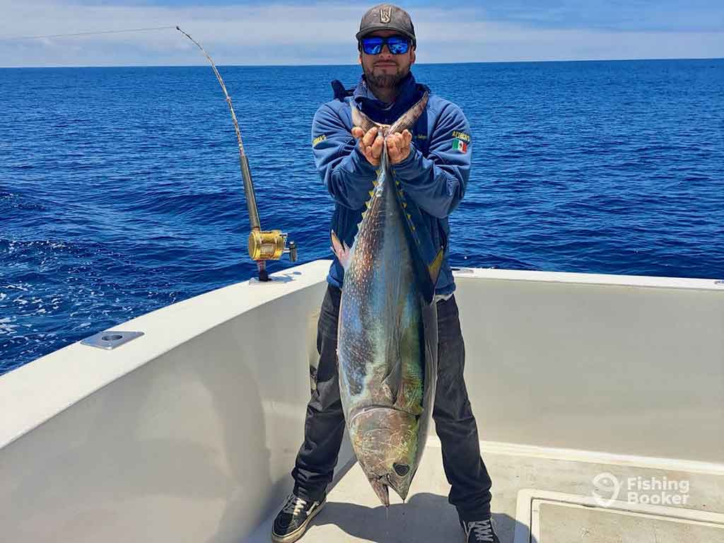 A man in sunglasses and a baseball cap standing on a fishing charter in Baja California and holding a large Tuna by its tail fin with the water behind him on a clear day