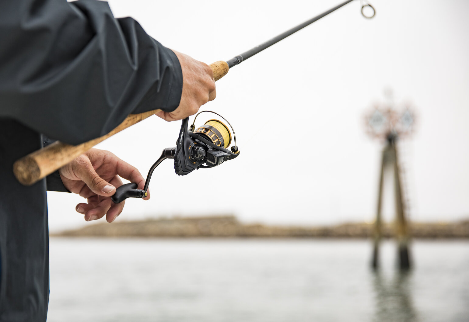Someone's holding and reeling a fishing rod by the water, with a navigational marker and shoreline in the background.