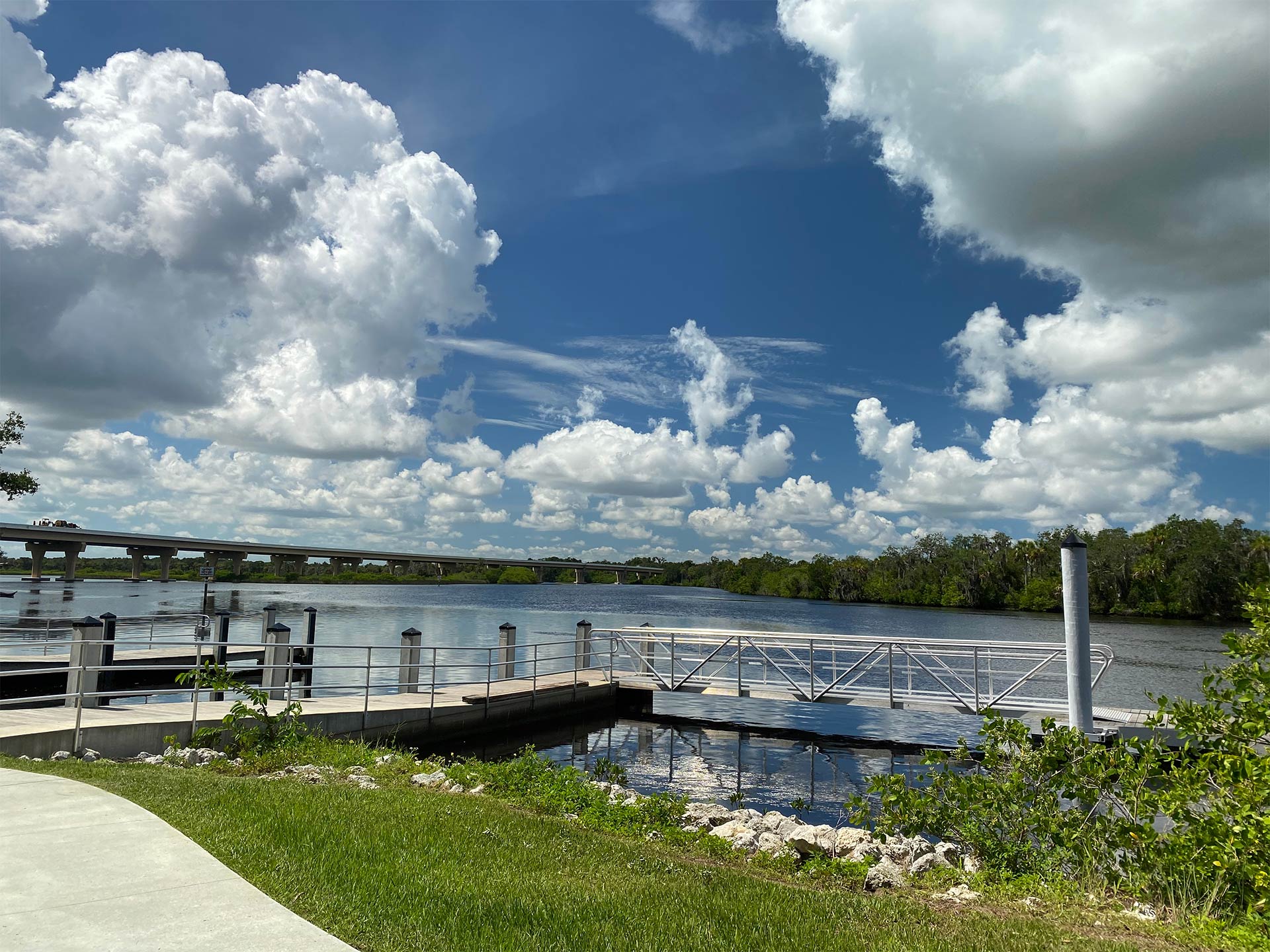 A view from a path of a boat dock in the Manatee River, with a bridge in the distance on a day of sunny intervals