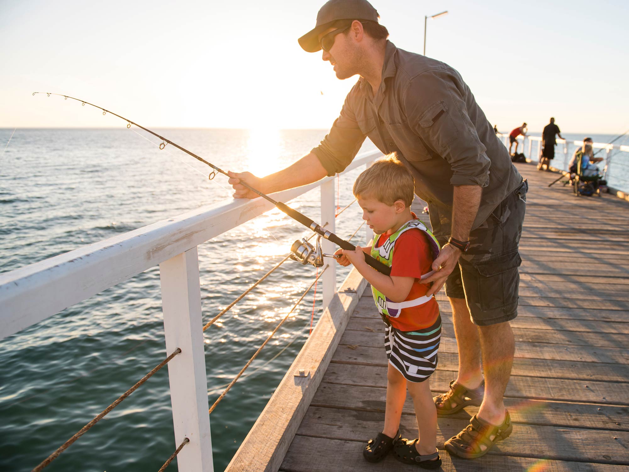 A father teaching his young son to fish on a fishing pier at sunset on a clear day