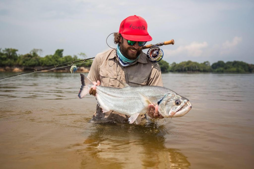 An angler standing in water with a fly rod and holding a Payara fish