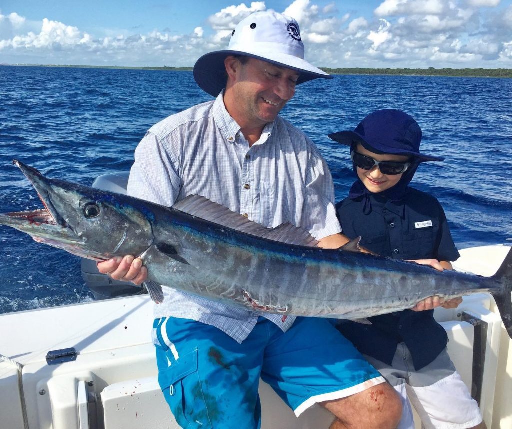 Father and son smiling and holding a giant Wahoo with water and skies as backdrop