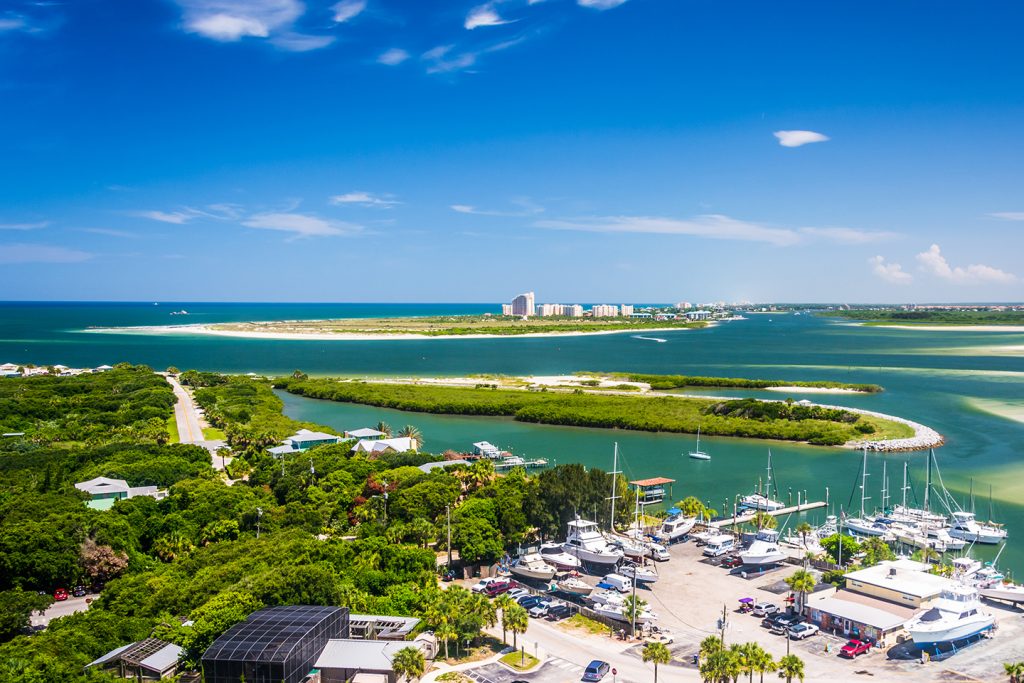 A charter fishing marina in New Smyrna Beach, FL, with boats out of the water in the bottom right, dense green trees on the left, and several green islands in the shallow sea in the center. Buildings are visible in the distance