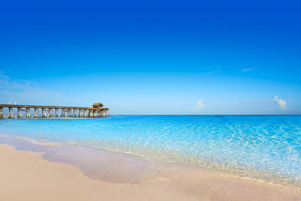 A sunny beach in Cape Canaveral, Florida. Yellow sand flows into bright blue water with an old fishing pier in the distance.
