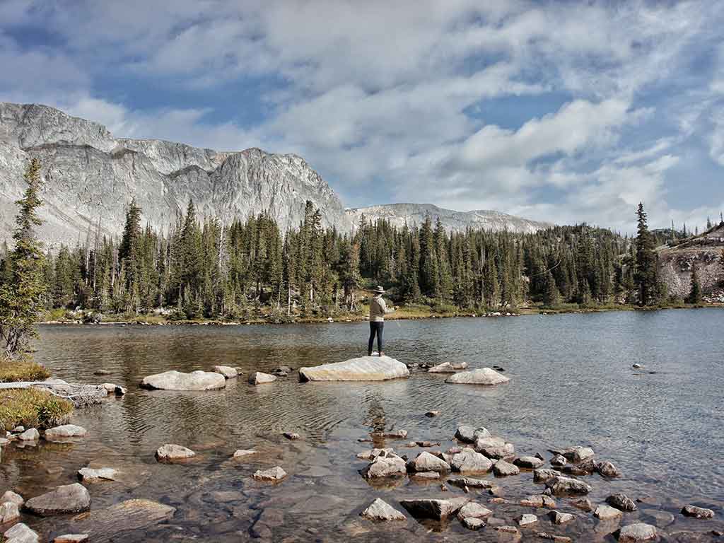 A view across the rocks in a waterway in North America towards a lone fly angler casting their line against an impressive backdrop of trees and a rocky mountain