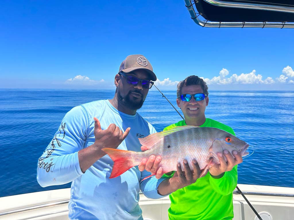 Two men on a charter boat posing for a photo while holding a beautiful Mutton Snapper they caught, with clear skies and calm ocean waters in the background.