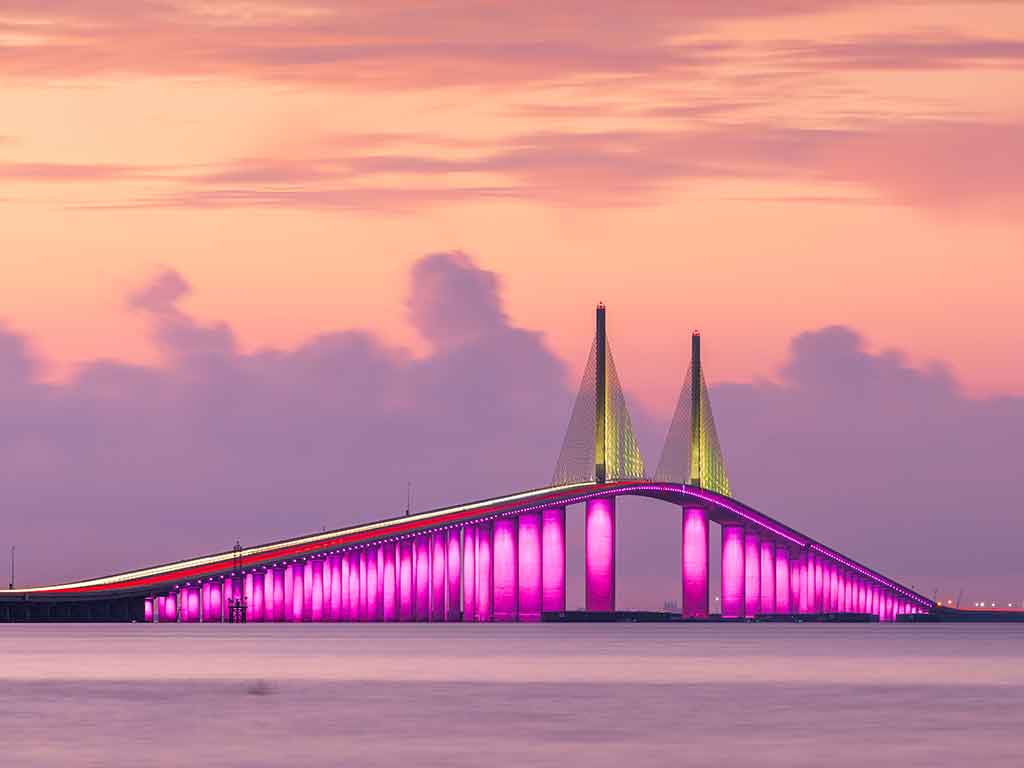 A viewa across the water towards the Sunshine Skuway Bridge in Tampa Bay at sunset, with the bridge's pink lights blending into the orange-red sky, with some clouds visible in the distance