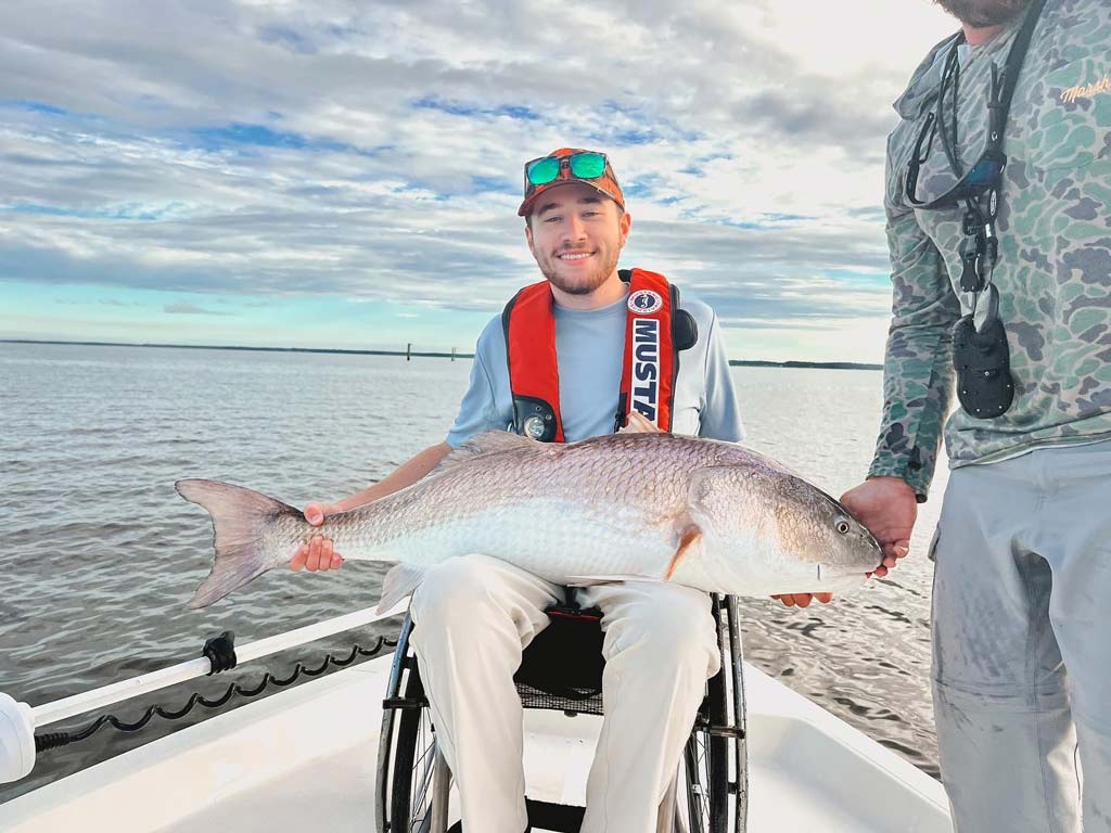 A smiling angler in a wheelchair on a boat, holding a huge Redfish in his lap, for which North Carolina holds the world record.