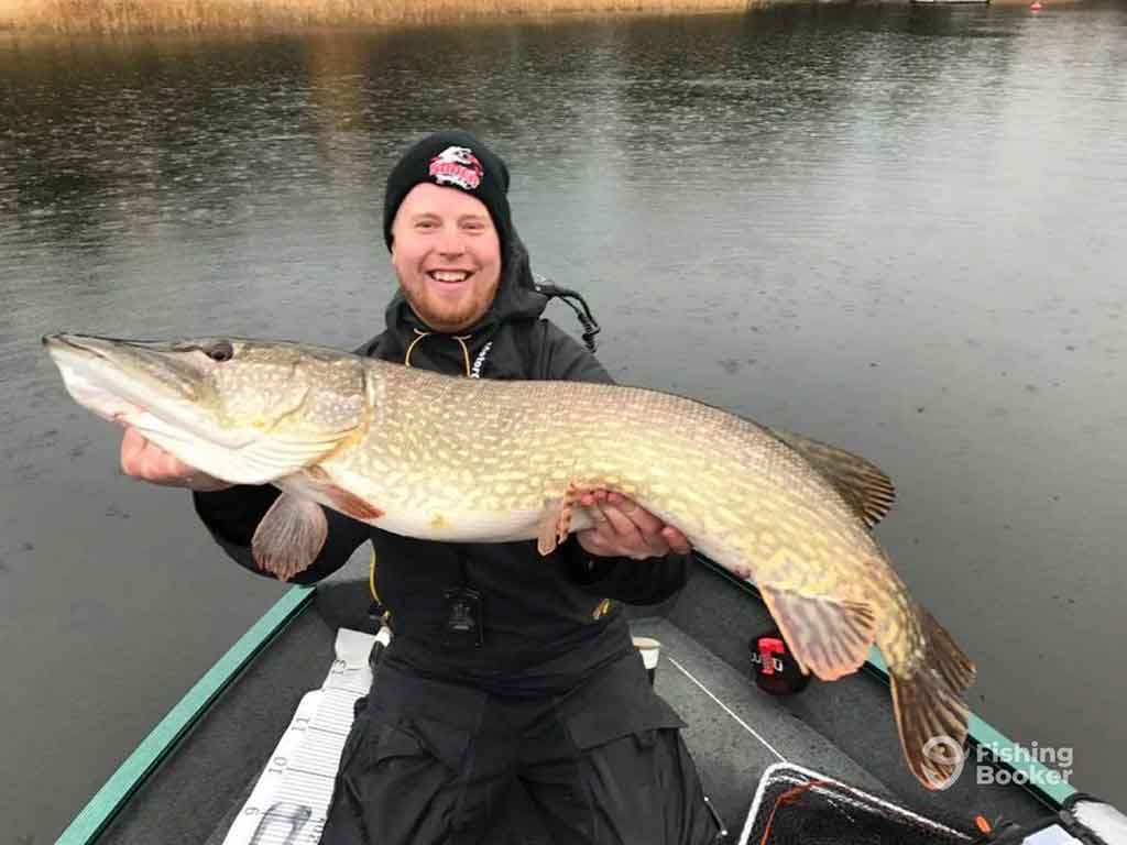 A young man in a beanie had on a rowing boat on a river in winter, holding a large Pike while smiling, with the waters visible around him