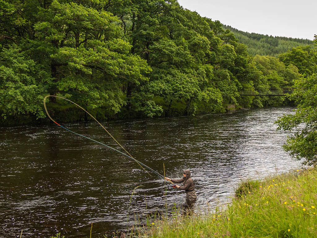 A view from atop a river bank looking down across the grass towards a fly angler, up to his waist in the water casting his line