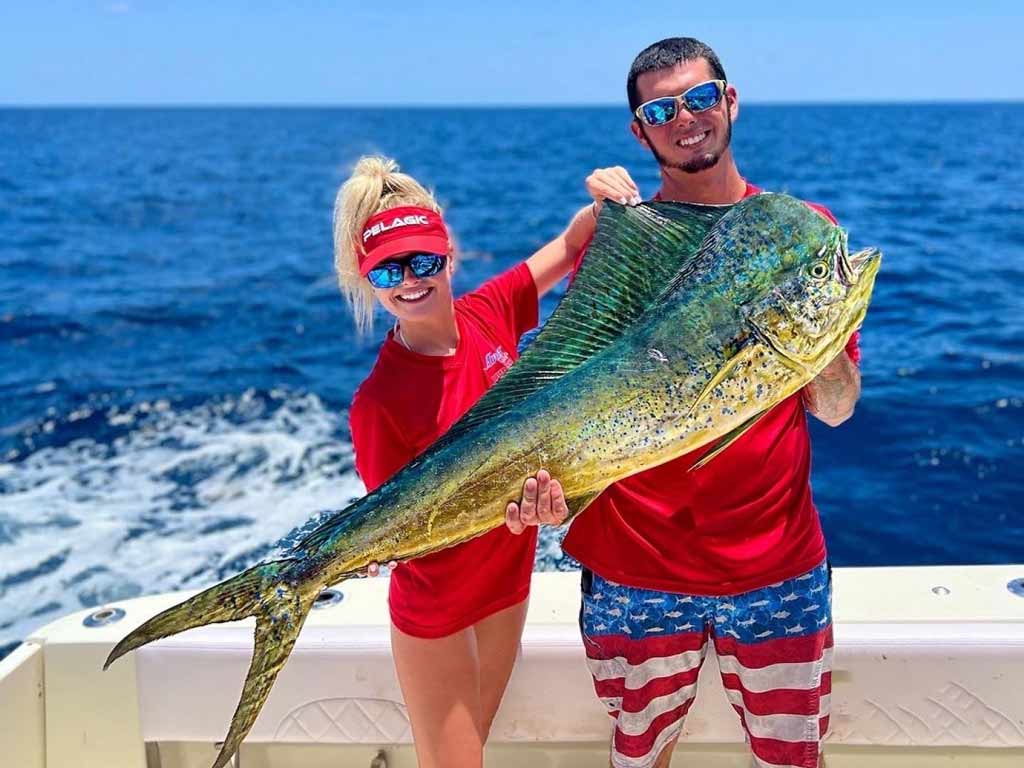 A photo depicting two anglers dressed up in patriotic patterns such as American colors and flag while standing on a deep sea fishing charter and posing with a big Mahi Mahi caught offshore in Florida.