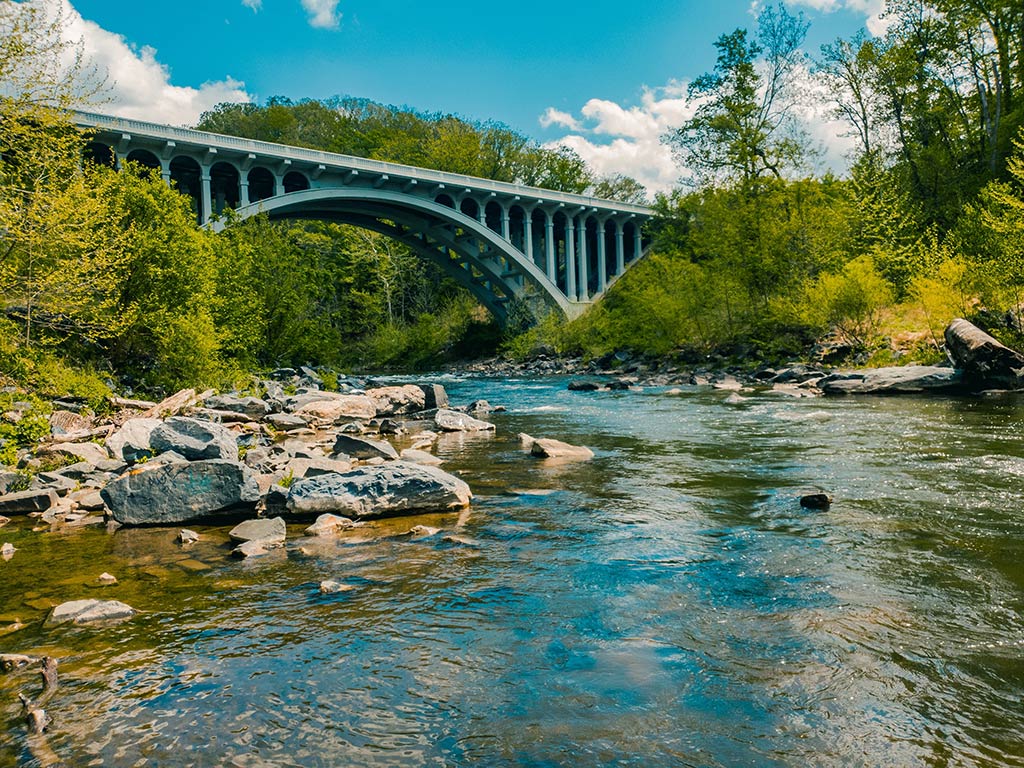 A view towards a small but high bridge crossing the Patapsco River near Baltimore on a clear day, with rocks visible in the foreground and greenery all around