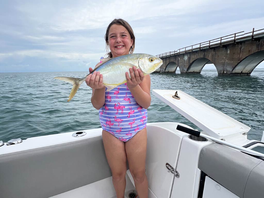 A smiling young girl in a flamingo swimsuit holds a Florida Yellow Jack while standing on a charter fishing boat with water and an arched bridge behind her.