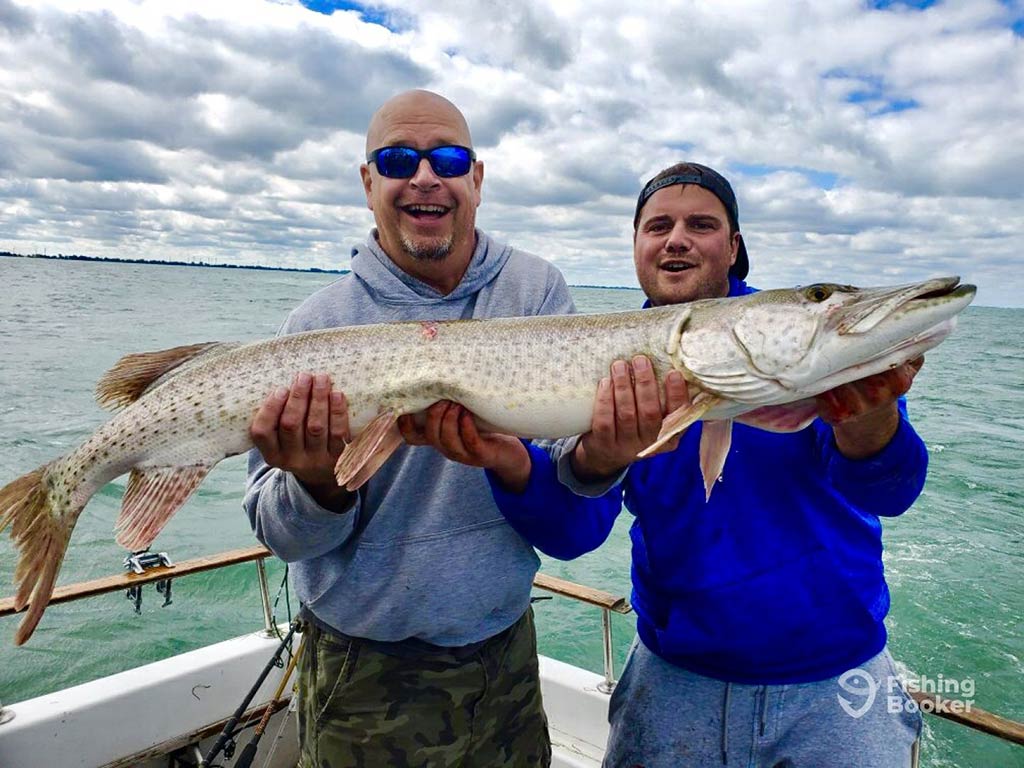 Two men on a fishing charter on a large lake holding a sizeable Pike on a cloudy day with the water visible behind them