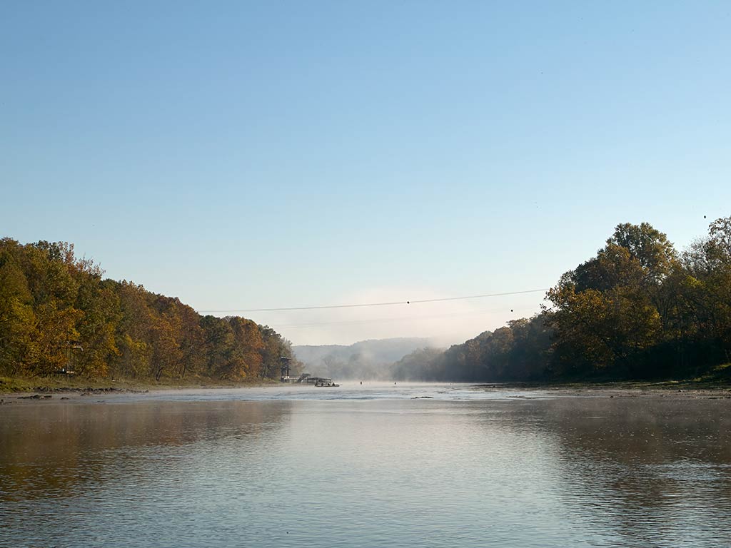 A view across the calm waters of the White River in Arkansas either first thing in the morning or last thing before dusk on a clear day, with large trees guarding the shoreline either side