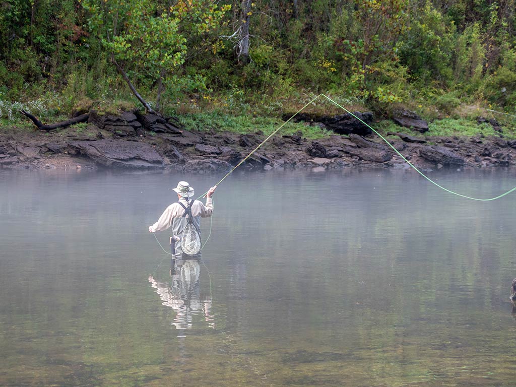 An angler wading up to his knees in a crystal clear river in Arkansas as he casts his fly fishing line