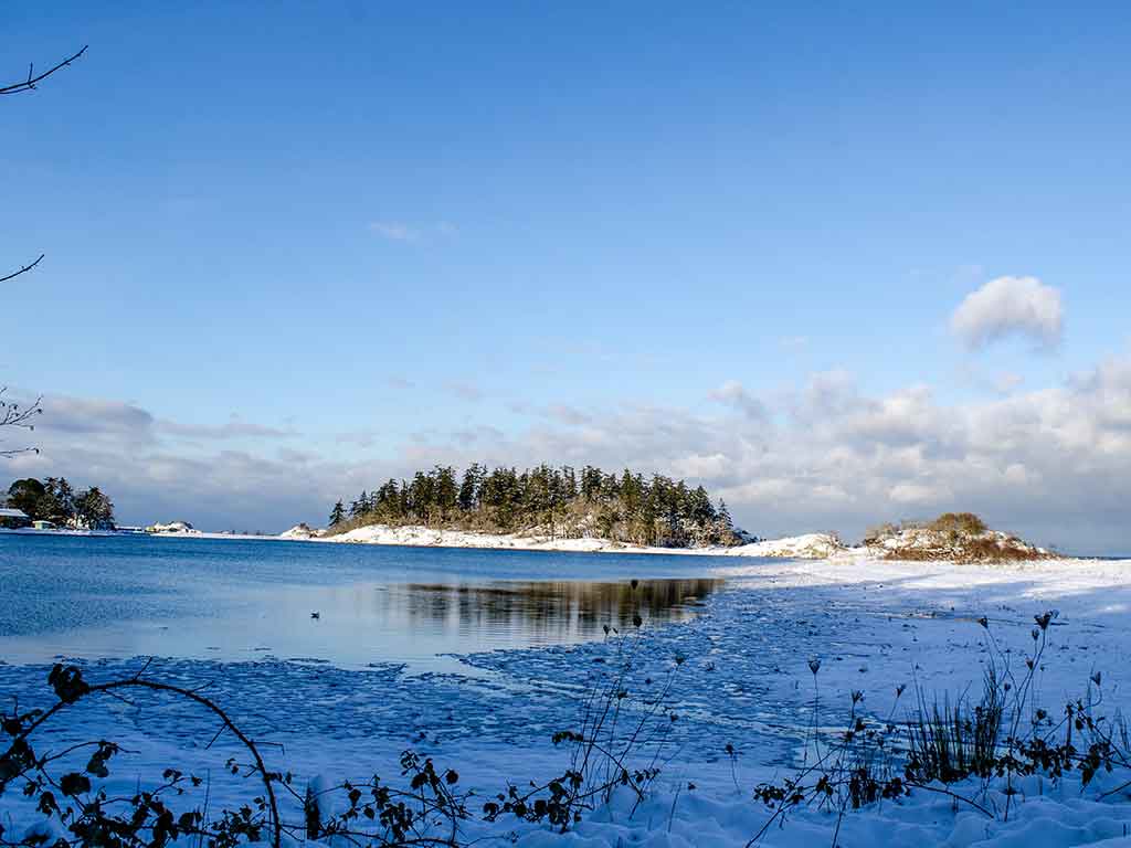 A view across the water near Nanaimo, BC, towards an island on a clear day, with snow visible on the ground across the area and the city visible in the distance on the left