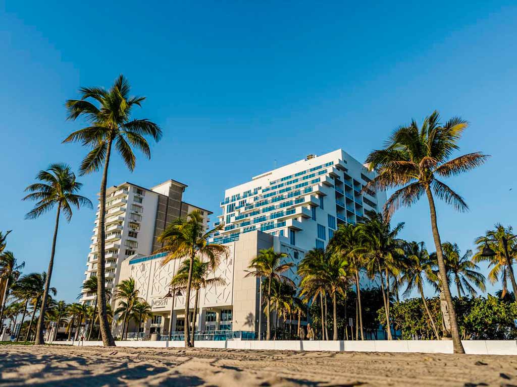 A photo of the exterior of Hotel Maren in Fort Lauderdale Beach, Florida, with palm trees sticking out of the sand in the forefront of the photo.