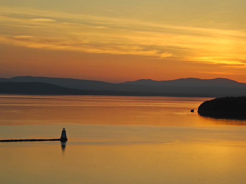 A view across the calm waters of Lake Champlain in Vermont at sunset, with the setting sun creating an orange hue across the sky