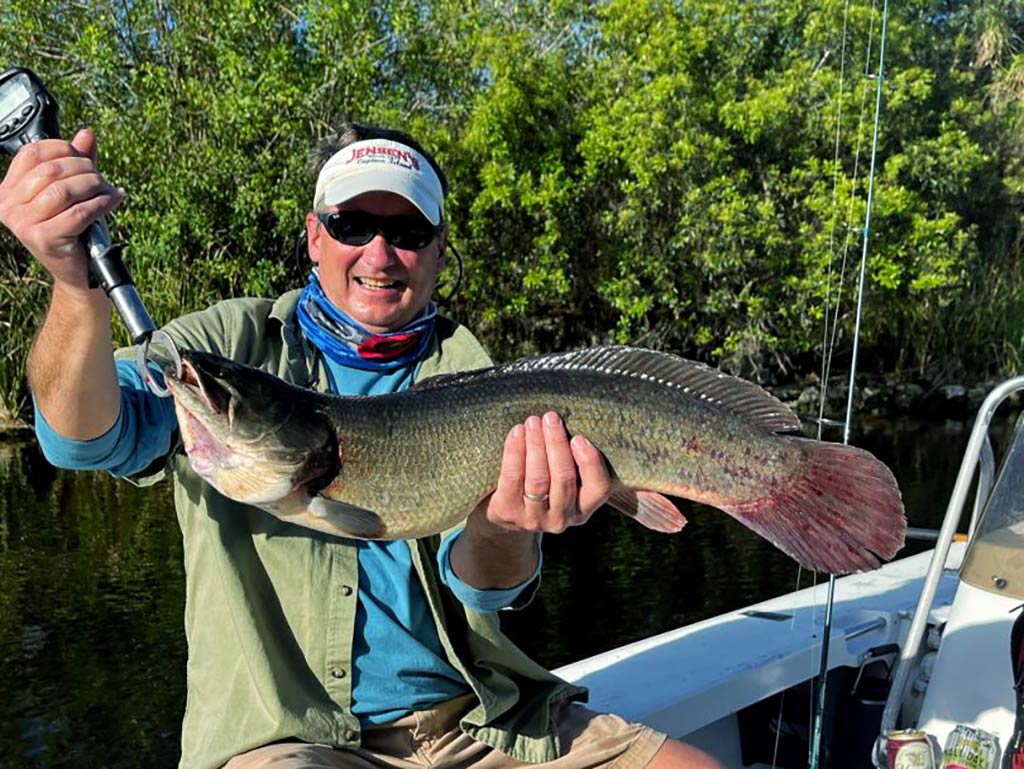 A man in a baseball cap and sunglasses smiles as he holds a sizeable Bowfin aboard a fishing boat in the Everglades during summer