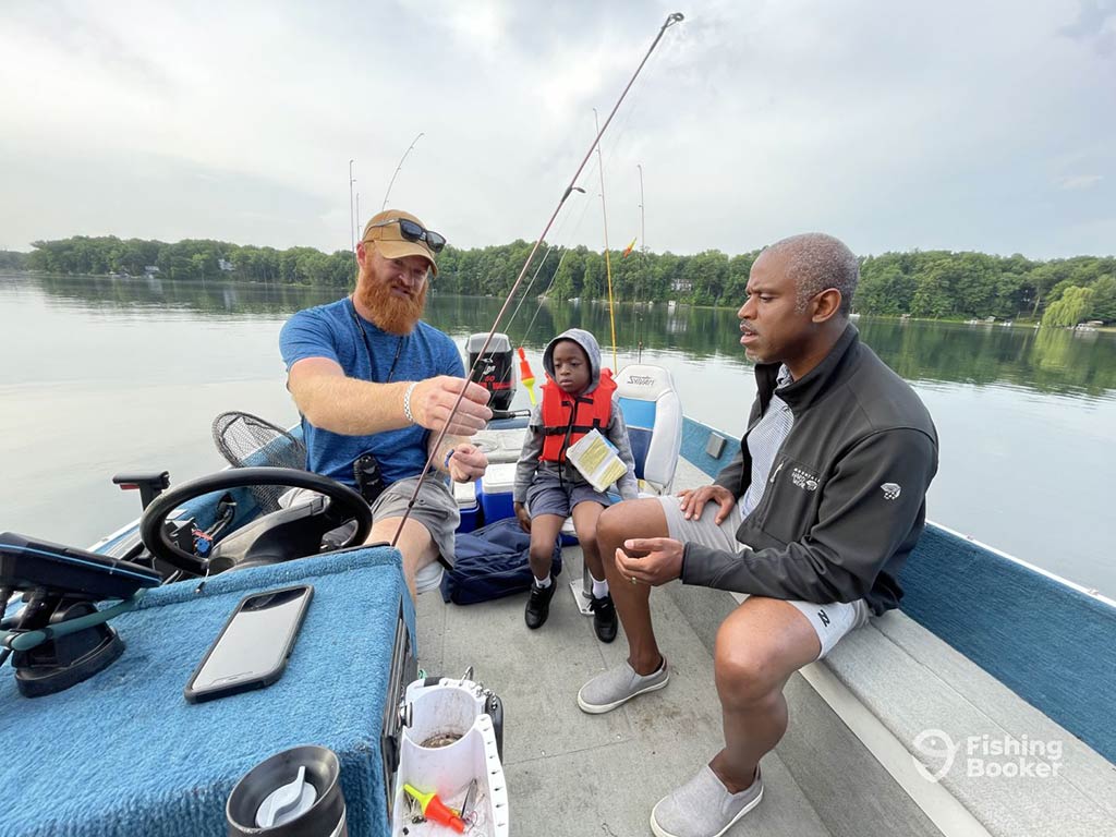 A man rigging and baiting a fishing line on a boat on a river as another man looks on and a child between them also looks