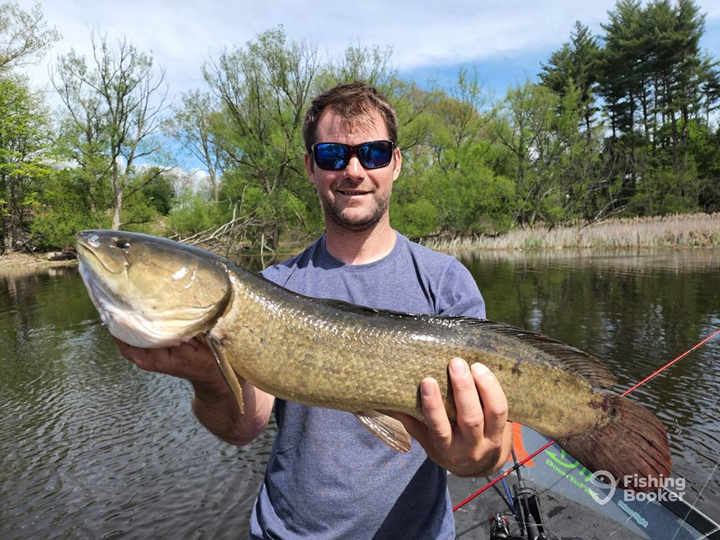 A man in sunglasses, standing on a fishing boat on a river and holdin a large yellow Bowfin on a clear day, with the water and some foliage visible behind him