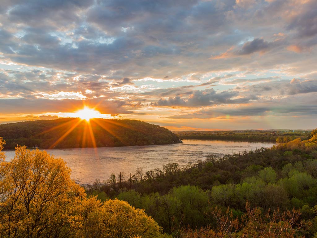 A stunning view of the sunset over Susquehanna River, with surrounding woods and slightly cloudy skies
