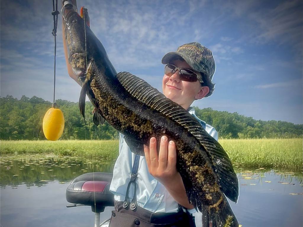 A teenage boy holding a sizeable Snakehead fish with a float hanging from its mouth against a background of calm waters and green shores